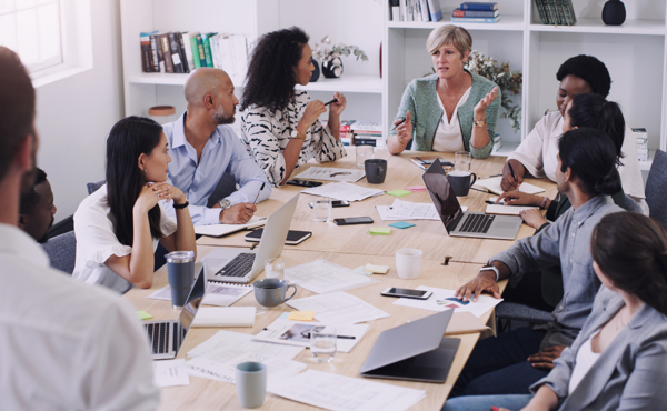 A group of people in business attire sit around a table in a modern office, discussing documents and laptops during a meeting.