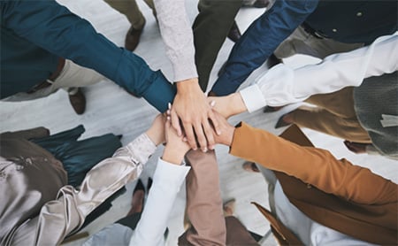 A diverse group of coworkers have their hands in the center of a huddle, signifying teamwork