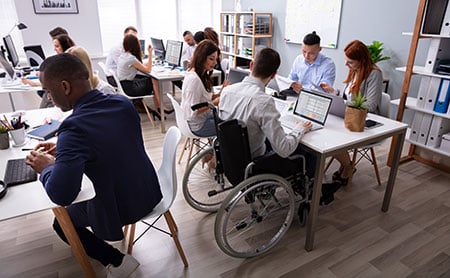 Diverse groups of young people studying on laptops around several tables, on person in a wheelchair