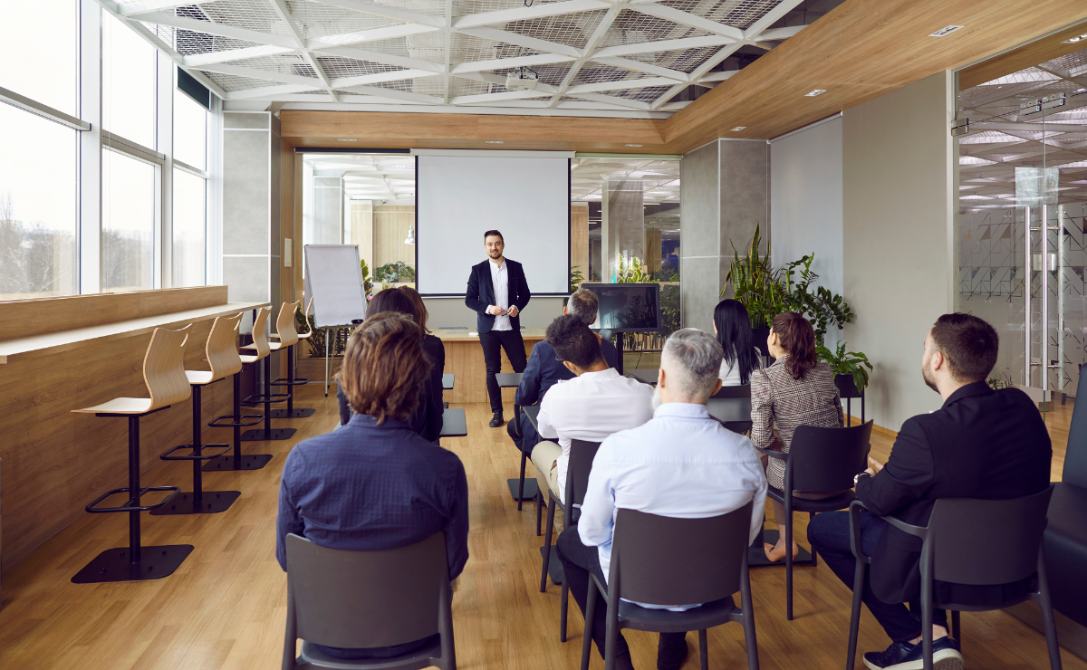 Man in front of video scren facing room of people sitting in chairs with their backs toward us in a modern room