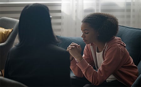 Adolescent girl on a couch holding hands together and looking pensive while talking to a counselor who's back is toward the camera