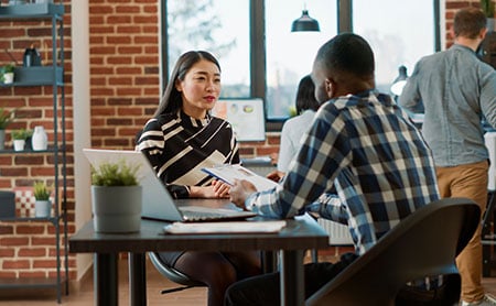 Two people discussing work at a table with a laptop and notepad in a modern office setting.