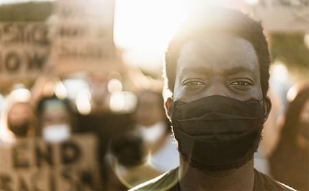 A Black man in a COVID-19 face mask, crowd from a racial equity protest visible behind him