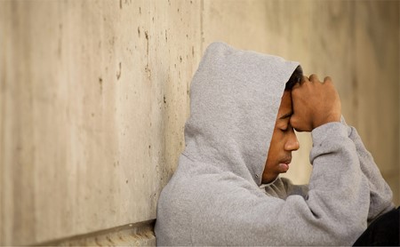 A Black teenage boy sits sadly, back pressed against a stone wall, head in hands