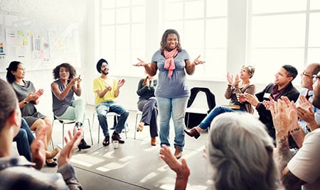 Black woman standing and talking in the center of a seated group applauding