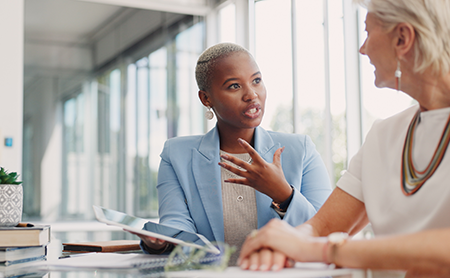 Two people sitting at a table with books and papers in front of them in a bright office setting having a conversation. 