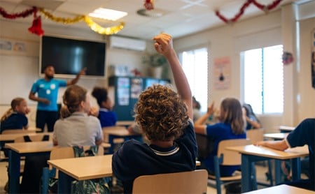 View of a classroom from the back, focus on a child with curly hair who has their hand raised