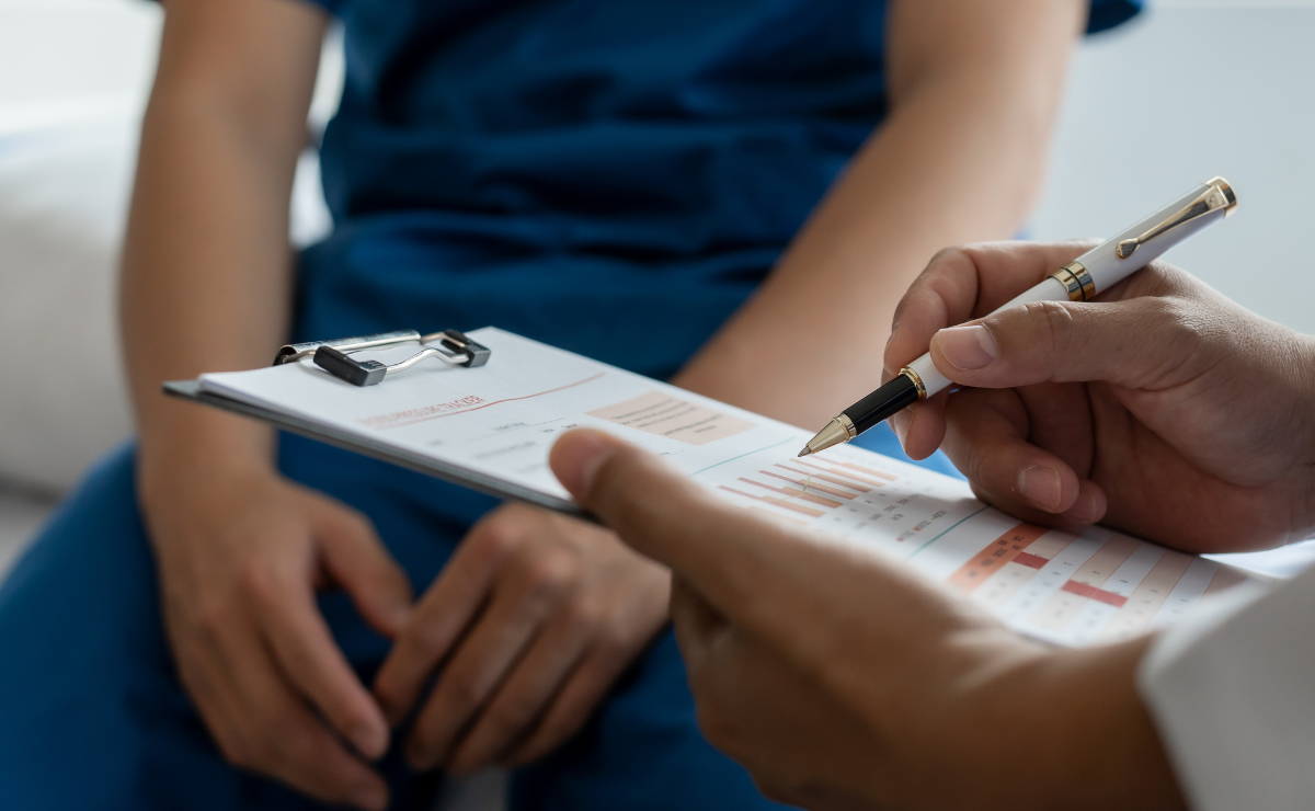 View from the clipboard as a doctor writes notes and woman sits in background during medical consultation.