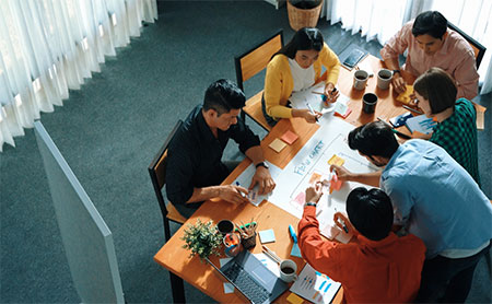 A group of individuals around a table in a meeting, discussing a flow-chart document, a laptop, and coffee cups are visible.