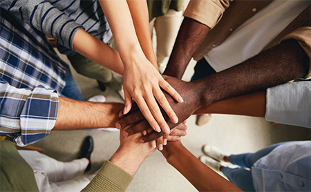 A diverse group of adults make a circle and each put a hand into the center, as a team-building cheer