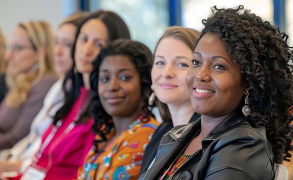 A diverse group of women are seated in the audience during a presentation, they have shifted to look at the off-camera speaker