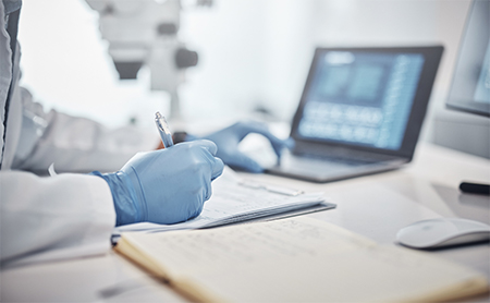 Midsection shot of a doctor, wearing blue medical gloves, takes notes from content on their laptop