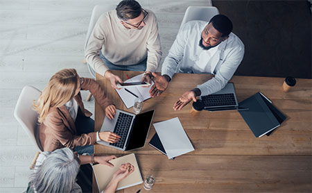 Four colleagues talking around a table with two open laptops, one person holding a tablet