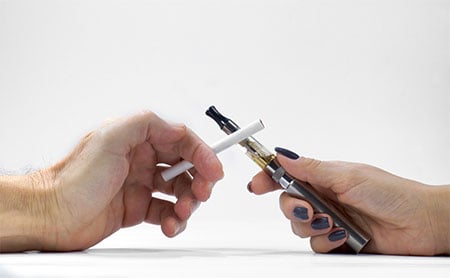 A man's hand with a cigarette and a woman's hand holding an e-cigarette. 