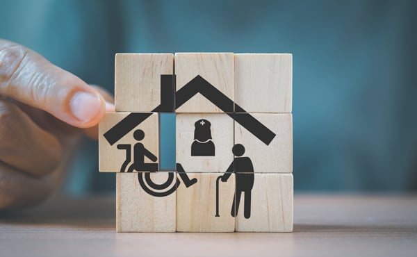 Hand completing wooden blocks with the design of a house containing symbols for a nurse, and two disabled people inside.