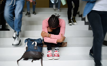 A high school student sits on busy steps, hugging her legs to her chest, face buried in her knees