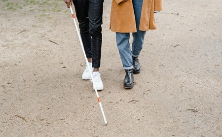 The legs of a blind person using a cane and their friend taking a walk on sandy ground.