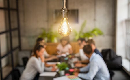 A close-up of a glowing light bulb hanging above a table with people in the background.