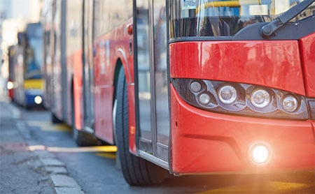 Three-quarter front view of a red city bus with another bus visible in the background.