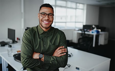 A Black man leans against a desk in his office and smiles broadly at the camera