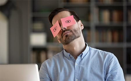Man sitting at his desk pretends to be awake by wearing Post-Its with open eyes drawn on