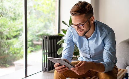 A smiling man sits on a bench in his living room, reading a tablet