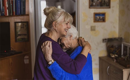 Two women embracing in a cozy room, surrounded by books and framed pictures.