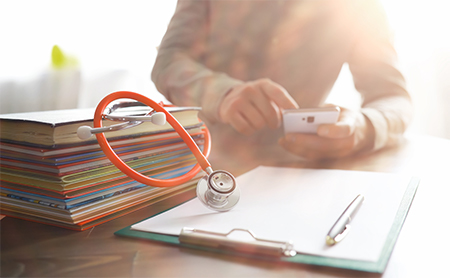 Midsection shot of a doctor sitting at her desk using her phone, in front of her are a pile of files, a notepad, and a stethoscope