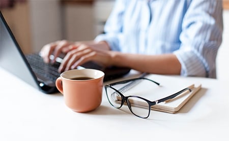 Midsection shot of a person working on a laptop at their desk, a cup of coffee and their eyeglasses next to them