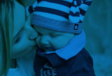 White woman with blond hair kissing a baby with a blue shirt and striped hat on the cheek