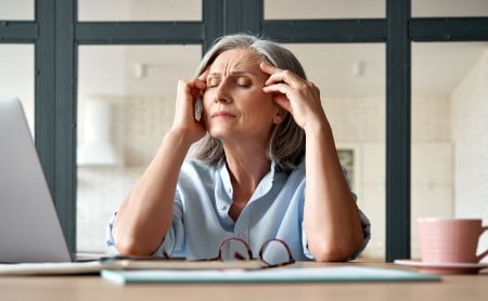 An older woman sits at her desk with her eyes closed, rubbing her temples, dealing with a terrible headache