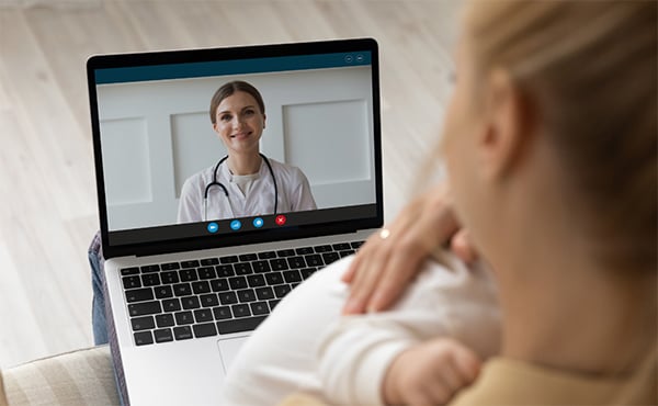 Over-the-shoulder shot of a mother holding her baby close during a telehealth appointment on her laptop