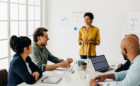 A woman leads a meeting in a bright office, speaking to colleagues seated at a table.