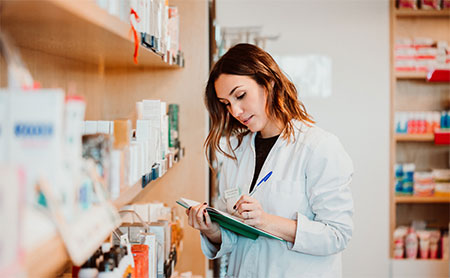 A pharmacist in a white coat writes on a clipboard with shelves of medications in the background.