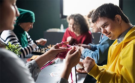 Closeup of a person teaching knitting to another with others playing chess in the background.