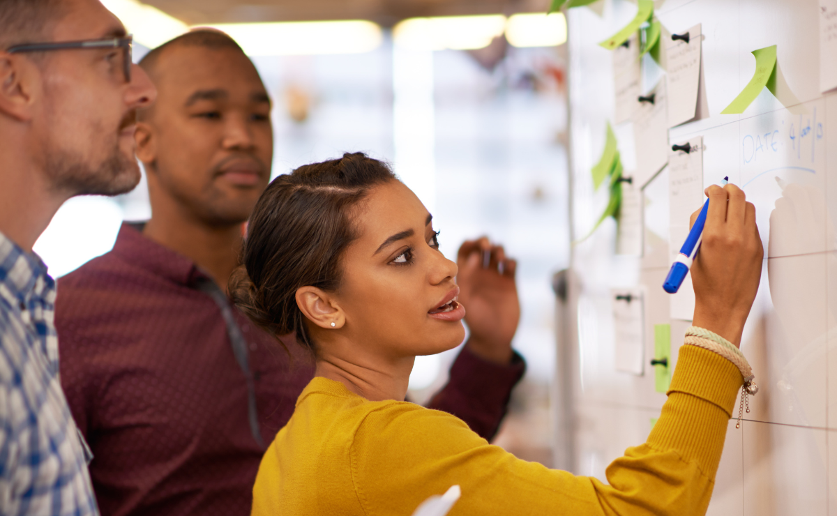 A team of three people collaborates around a whiteboard, actively discussing and writing down their ideas and plans.