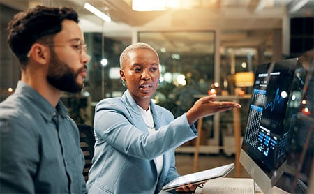 Two people in an office looking at data charts on a computer monitor.