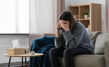 A depressed woman sits on the couch making a phone call, a box of tissues, glass of water, and overturned bottle of pills in front of her.