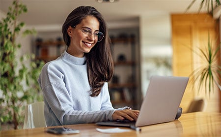 Smiling woman wearing glasses typing on a laptop in a home setting with plants