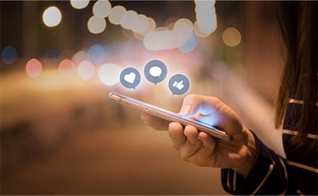 Midsection shot of a woman's hand using a smartphone with social media icons above phone. Blurred street in background. 