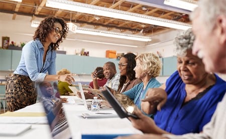 A group of middle aged people sit at a folding table and work on laptops while speaking with a young woman